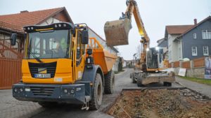 Ein Mobilbagger hält seine Schaufel über einem gelben Baustellenkipper neben der aufgegrabenen Fahrbahn einer Wohnstraße.