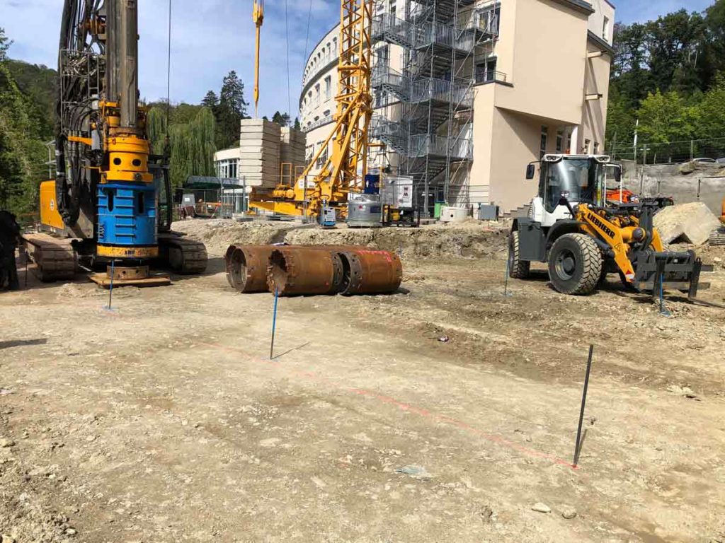 Bohrgerät, Radlader und abgelegte Stahlrohre auf einer Baustelle vor einem eingerüsteten Gebäude.