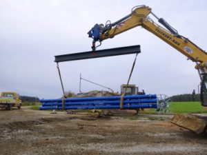 Ein Bagger hebt ein Bündel blauer Rohre mit einer Stahltraverse und Hebegurten auf einer Baustelle.