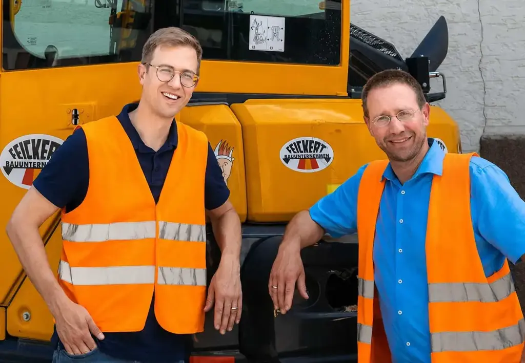 Zwei Männer in orangefarbenen Warnwesten lächeln vor einer gelben Baumaschine.