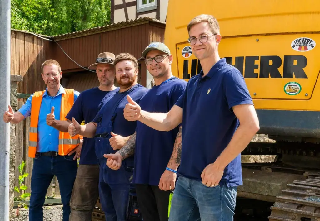 Fünf Männer posieren mit erhobenen Daumen vor einem gelben Raupenbagger.