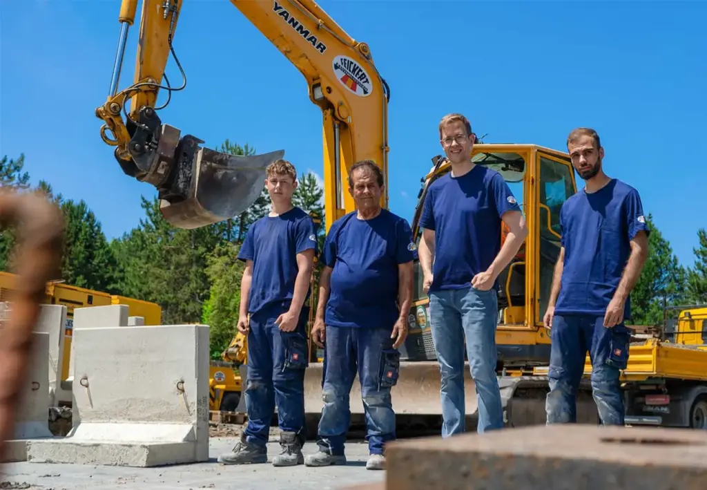 Vier Männer in blauer Arbeitskleidung stehen neben Betonfertigteilen vor einem gelben Bagger.
