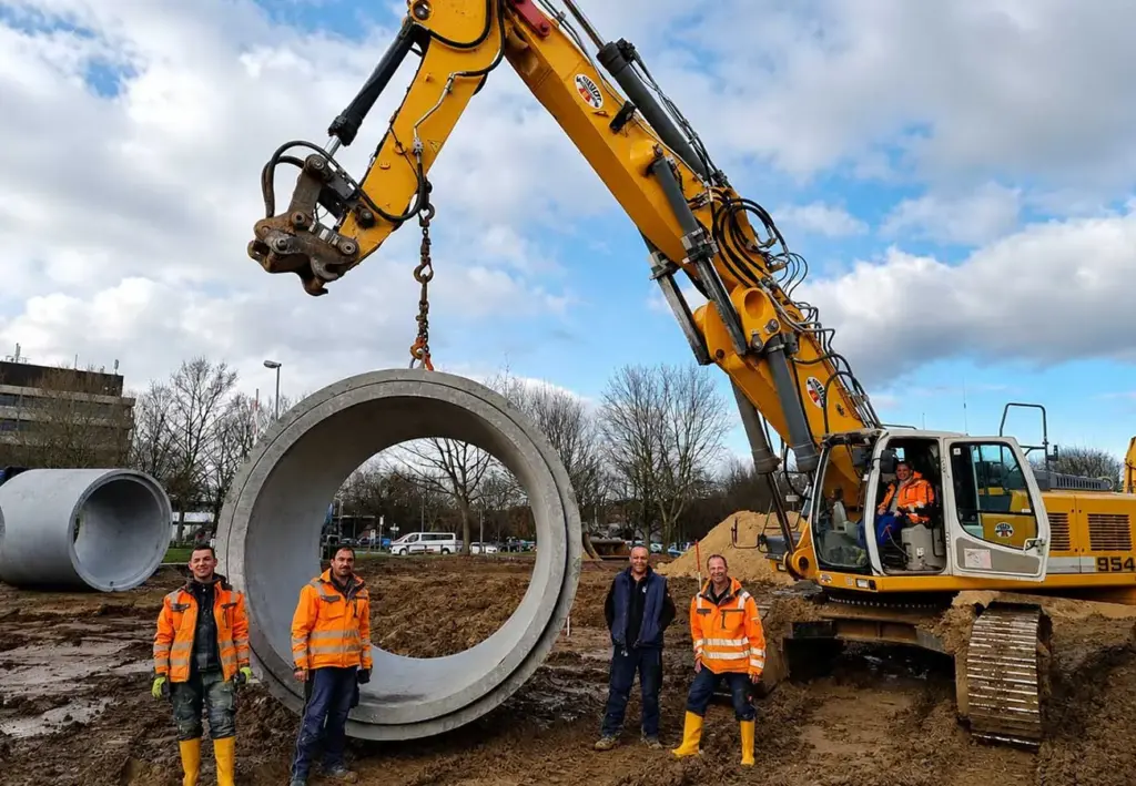 Vier Männer posieren neben einem großen Betonrohr, das an einer Kette am Ausleger eines Raupenbaggers hängt.