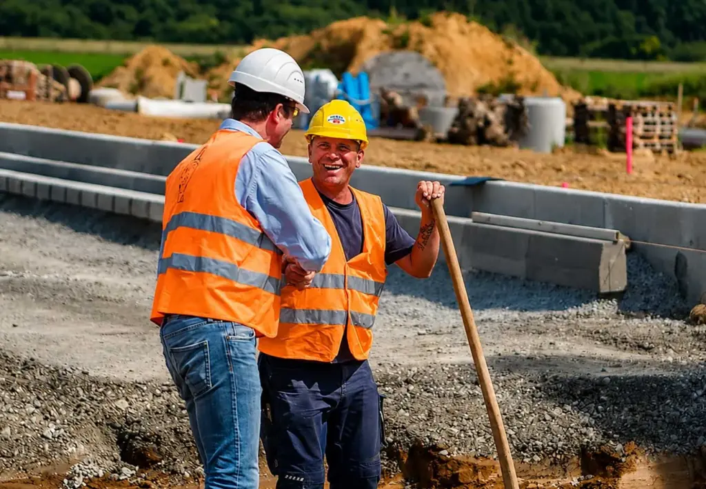Zwei Bauarbeiter mit Schutzhelmen und Warnwesten unterhalten sich auf einer Baustelle; einer lächelt und hält einen langen Werkzeugstiel.