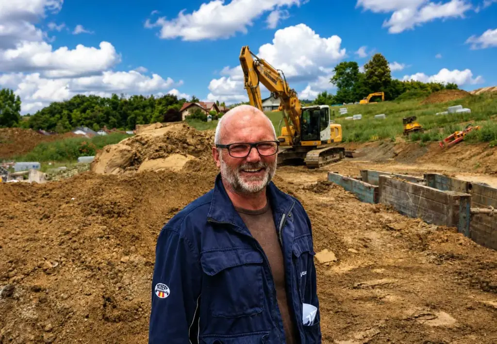 Ein Mann mit Brille und grauem Bart lächelt auf einer Baustelle mit Erdhaufen und einem Bagger im Hintergrund.