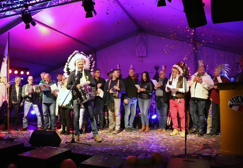 Trommler mit Federschmuck vor einer singenden Gruppe auf der violett beleuchteten Bühne im Festzelt.